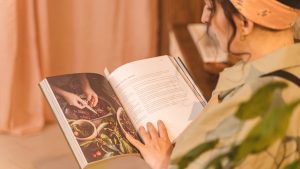 A woman reading a cookbook with colorful images and recipes in a warm indoor setting.