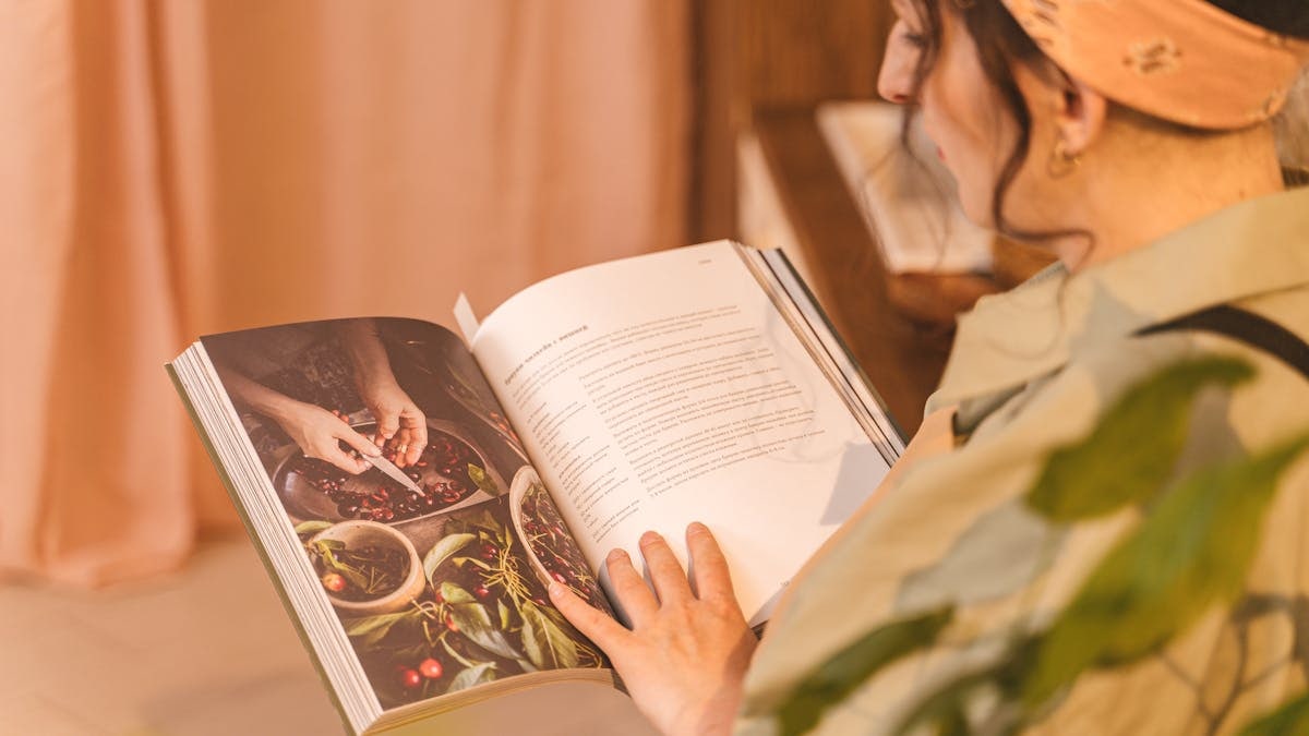 A woman reading a cookbook with colorful images and recipes in a warm indoor setting.